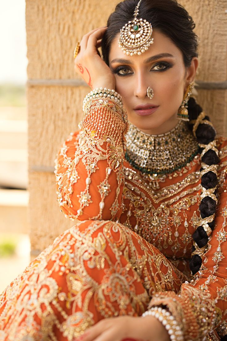 a woman in an orange and gold outfit with jewelry on her head, sitting against a wall