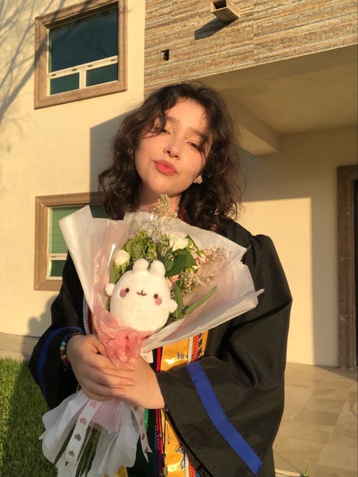 a woman holding flowers and a stuffed animal in front of a building with grass on the ground