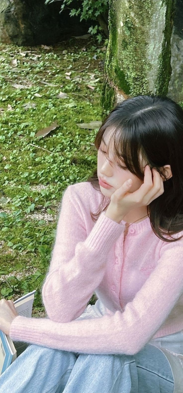 a young woman sitting on the ground with her hand under her chin, reading a book