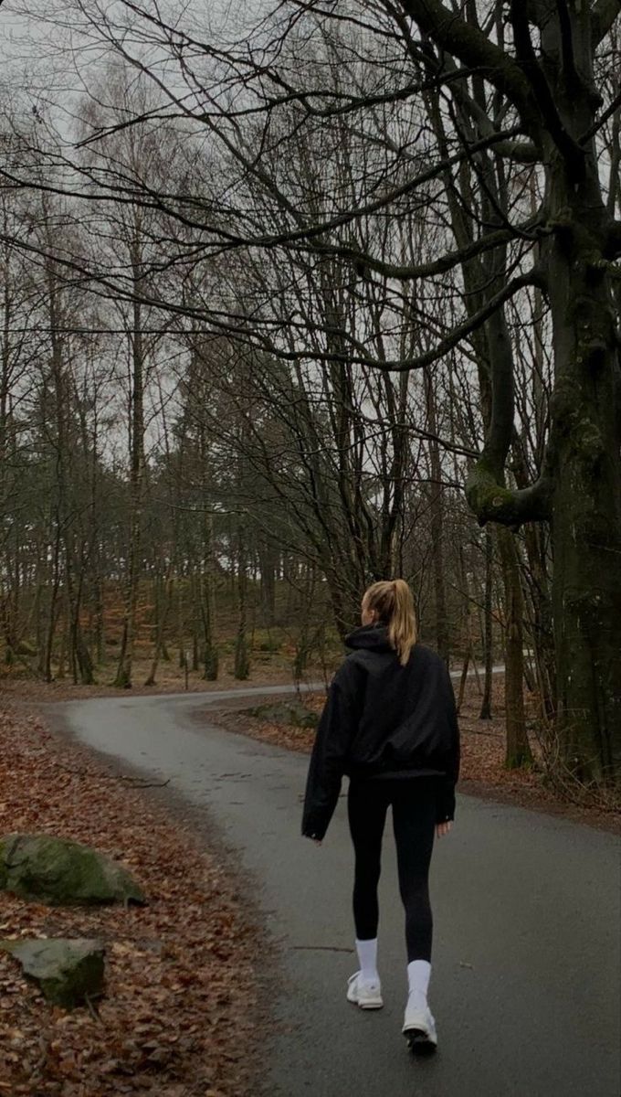 a woman walking down a road in the middle of a forest with no leaves on it