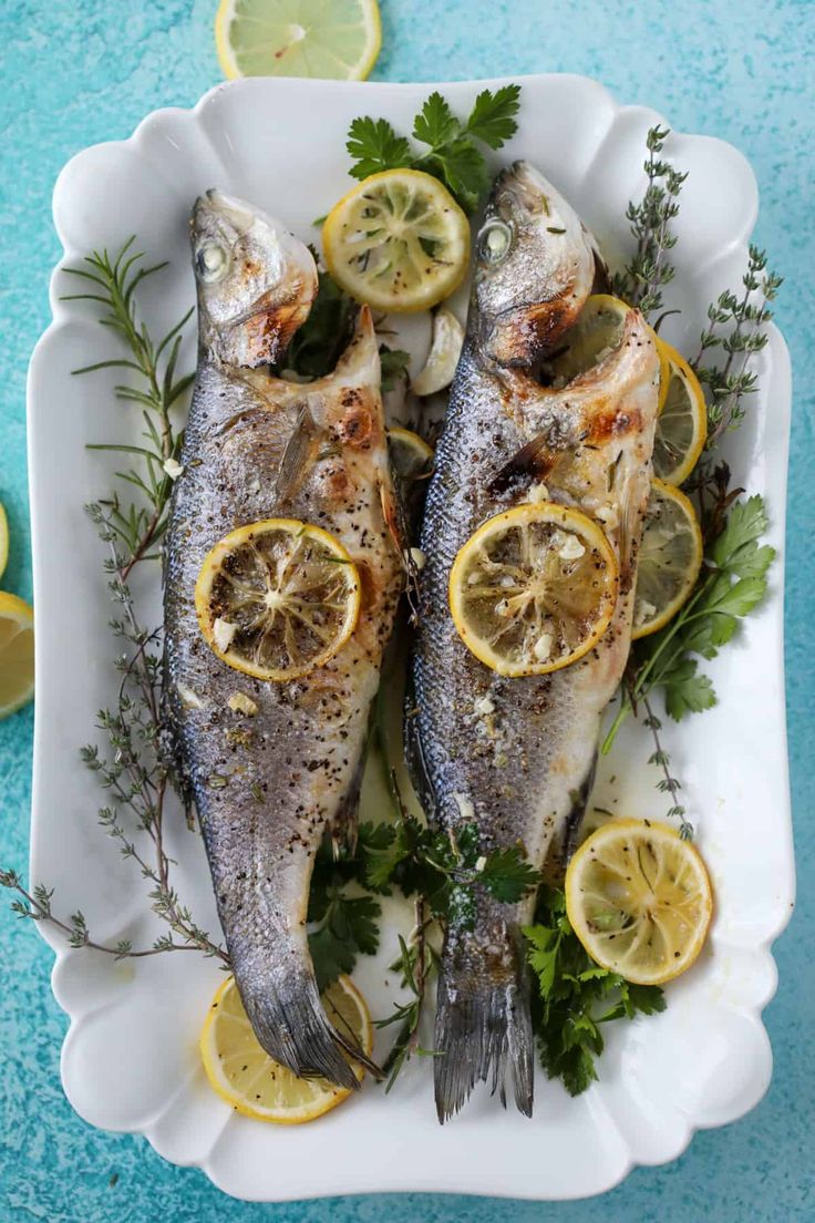 two fish with lemons and herbs on a white platter next to sliced lemons