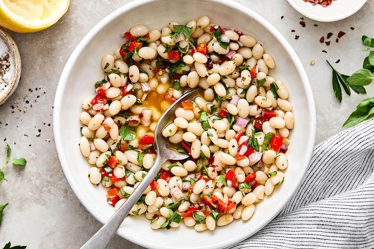 a white bowl filled with bean salad next to lemons and parsley on the side