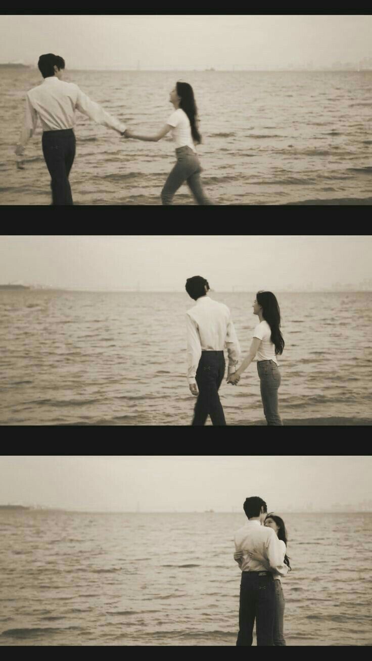 two people holding hands while standing on the beach next to the ocean with their arms around each other