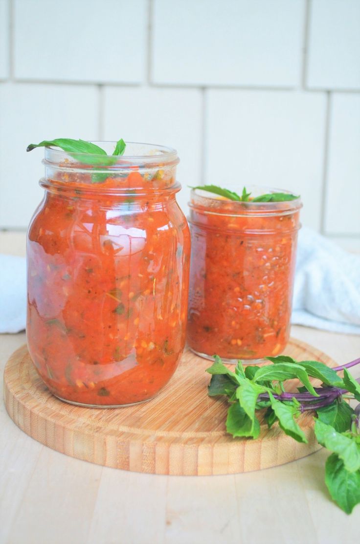 two jars filled with red sauce sitting on top of a wooden cutting board next to green leaves