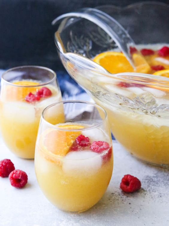 three glasses filled with lemonade and raspberries next to a bowl of oranges