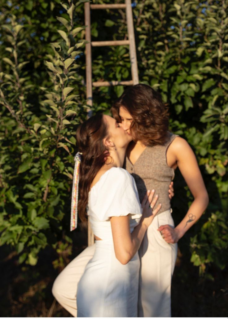 two women are kissing each other in front of some bushes and trees with a ladder behind them