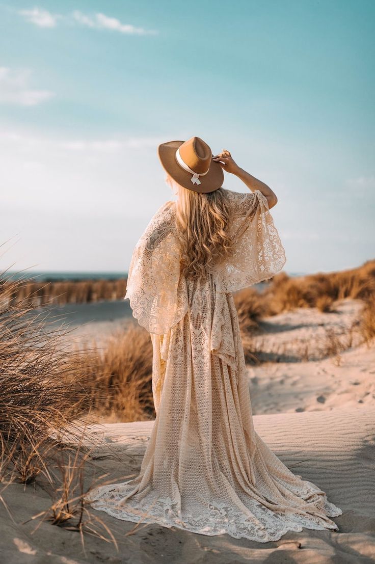 a woman in a dress and hat standing on the sand dunes looking out at the ocean