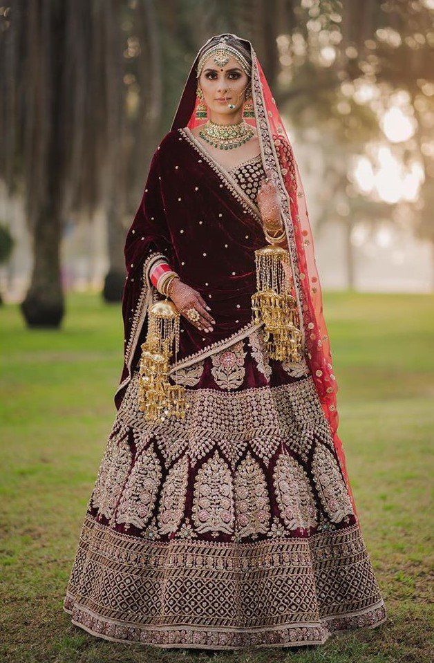 a woman in a red and gold bridal outfit standing on grass with trees behind her