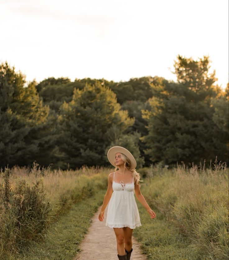 a woman in a white dress and hat walking down a dirt road