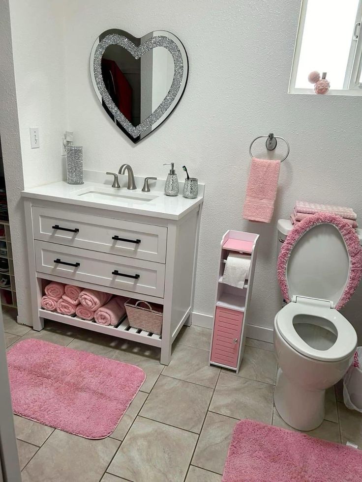 a white bathroom with pink rugs on the floor and toilet in the corner, along with a heart shaped mirror over the sink