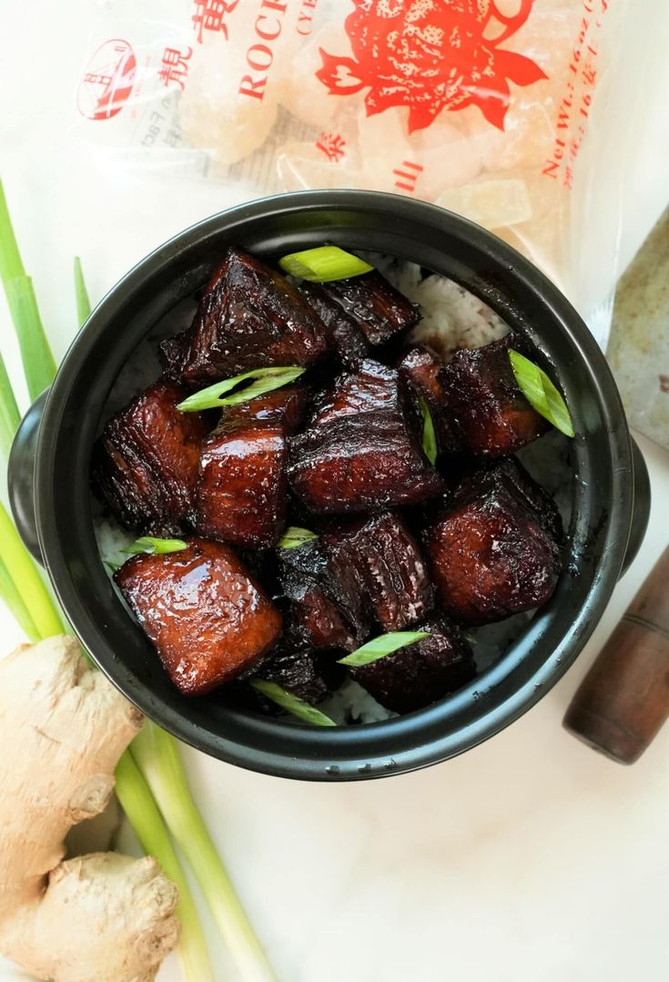 a black bowl filled with cooked food next to some green onions and ginger sticks on top of a white table