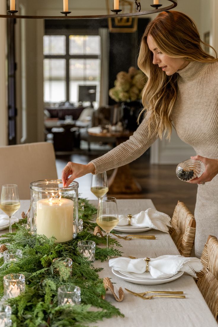 a woman setting a table with candles and greenery on the dining room table in front of her