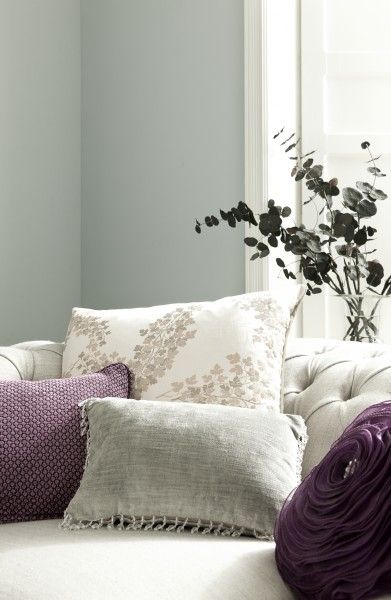 a white couch with purple pillows on it and a vase full of flowers in the corner