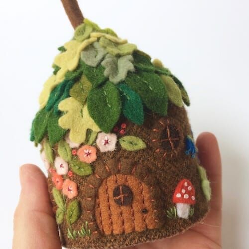 a hand holding a small felt house with flowers and leaves on the outside, in front of a white background