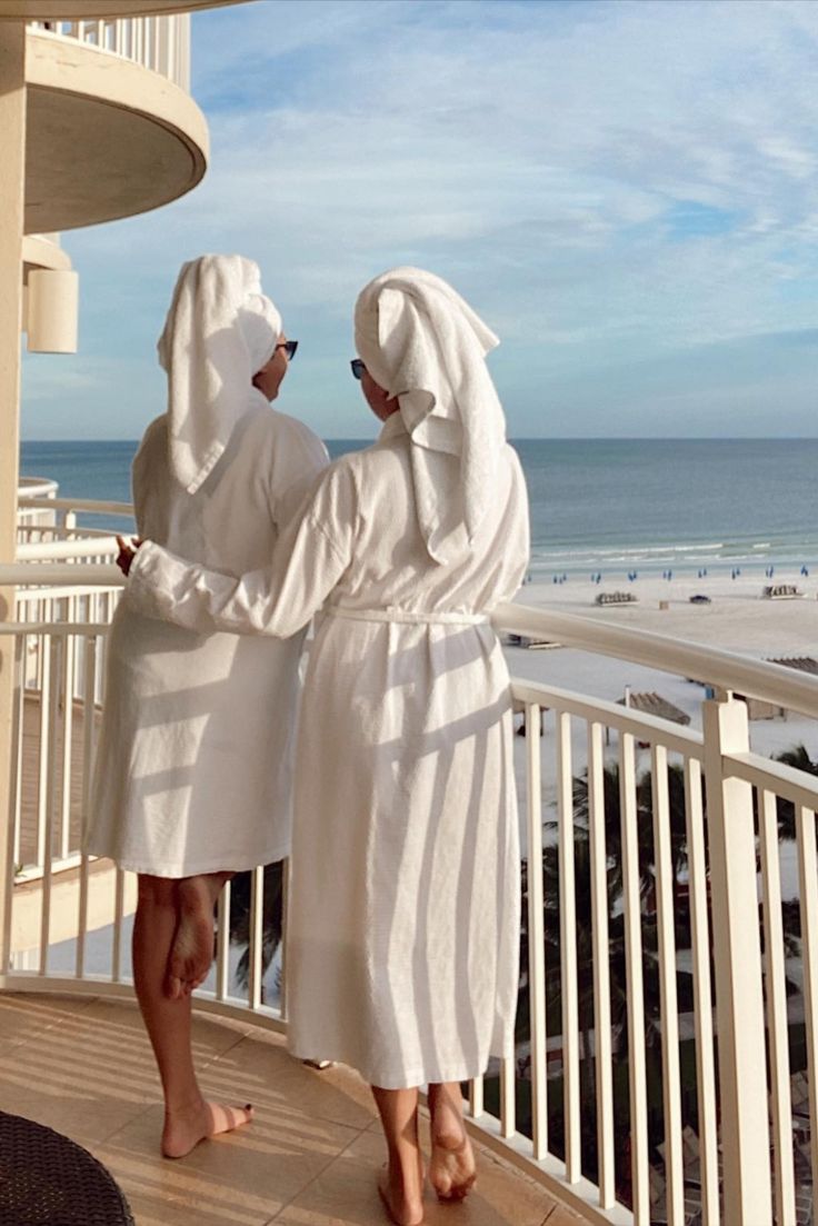 two people in robes standing on a balcony looking out at the beach and ocean,