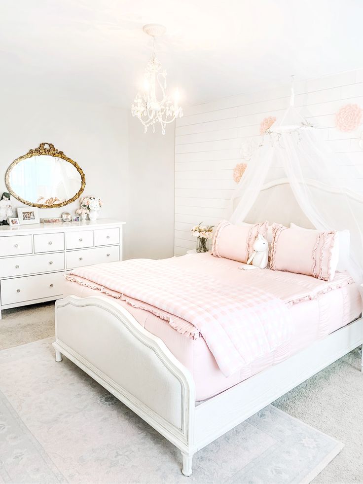 a white bed with pink comforter and pillows in a small room next to a chandelier