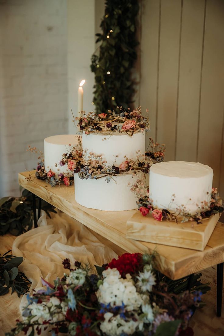 three white cakes sitting on top of a table covered in flowers and greenery next to a lit candle