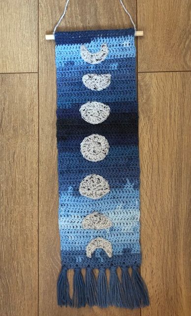 a crocheted wall hanging with three moon and clouds on it, in the middle of a wood floor