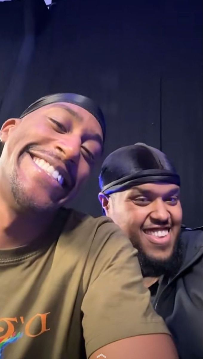 two men are smiling and one is wearing a hat with his head turned to the side