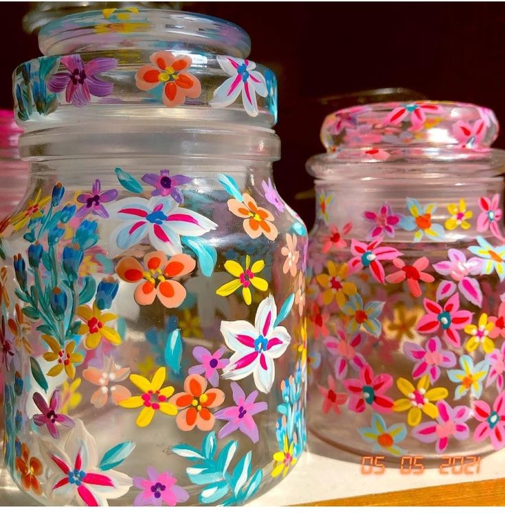 three jars with painted flowers on them are sitting on a counter top, one is clear and the other is bright