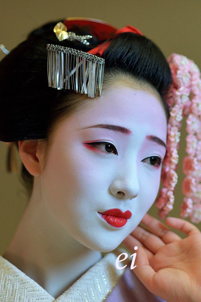 a geisha woman with white makeup and red hair holding her hand near her face