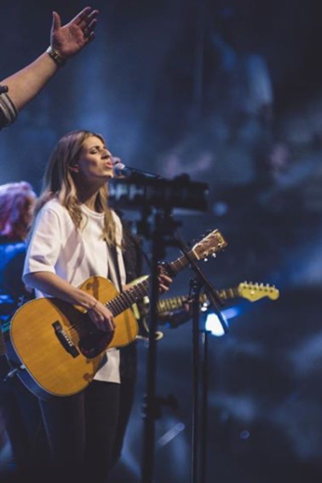 a woman singing into a microphone while holding a guitar and standing next to a man