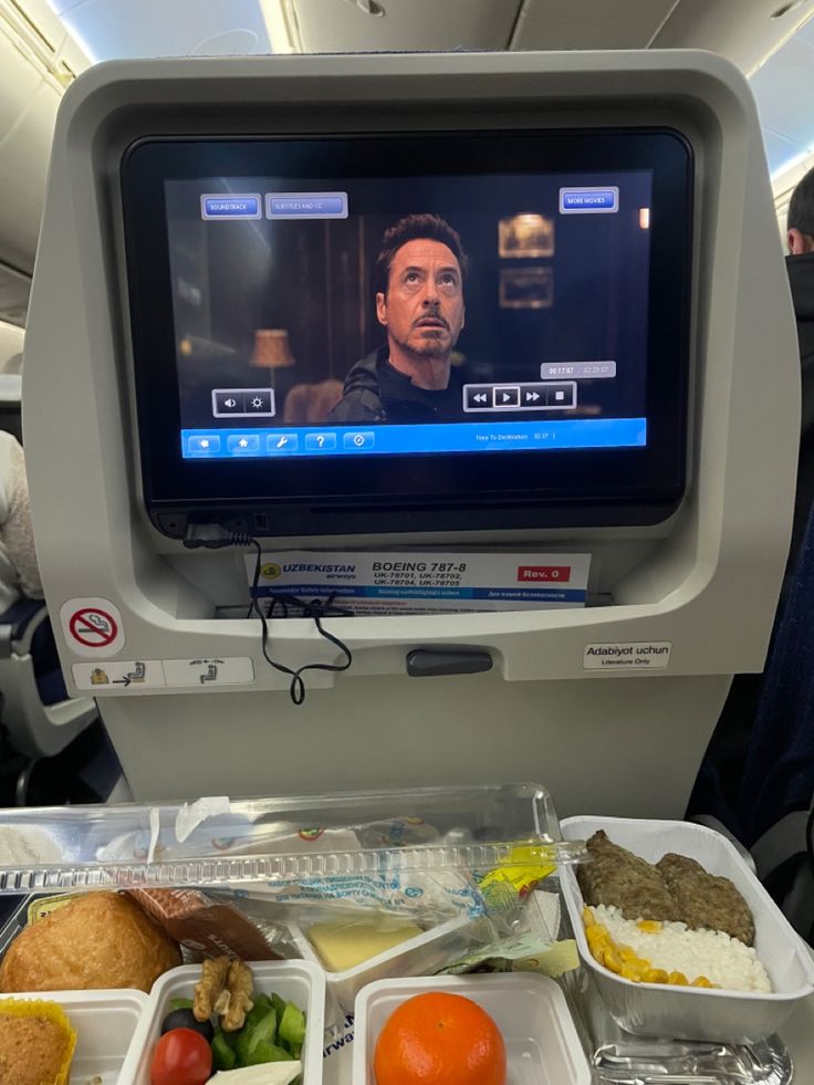 an airplane seat with food on it and a tv screen in the back ground,