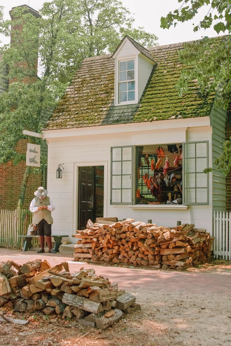 a man standing in front of a pile of wood next to a white house with green shingles