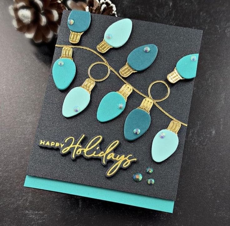 a happy holidays card with blue and gold ornaments on the front, sitting on a table next to a pine cone