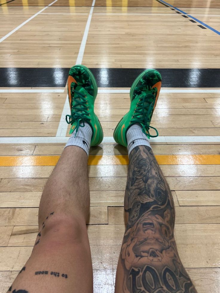 a man with tattoos on his legs and green shoes is sitting in the middle of a basketball court