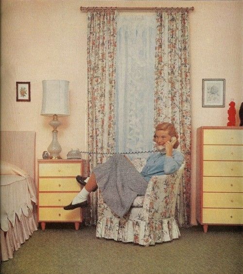 a woman sitting in a chair talking on a cell phone next to a dresser and bed