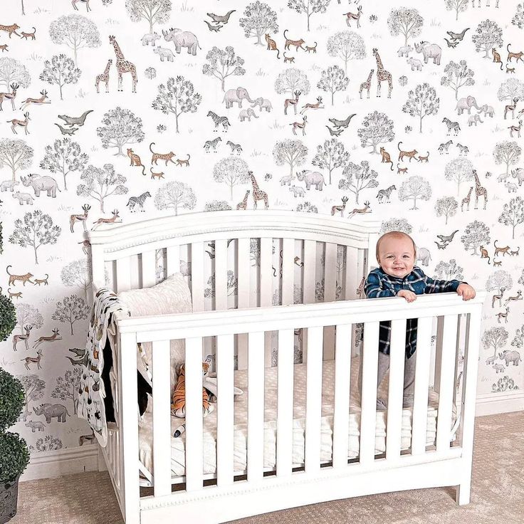 a baby sitting in a white crib next to a wall with animals on it