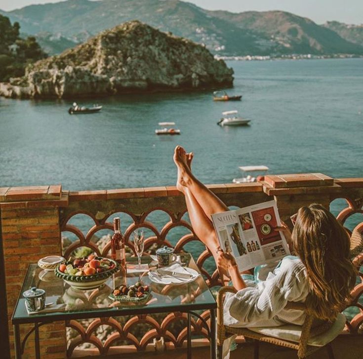 a woman sitting at a table reading a magazine overlooking the ocean and boats in the water