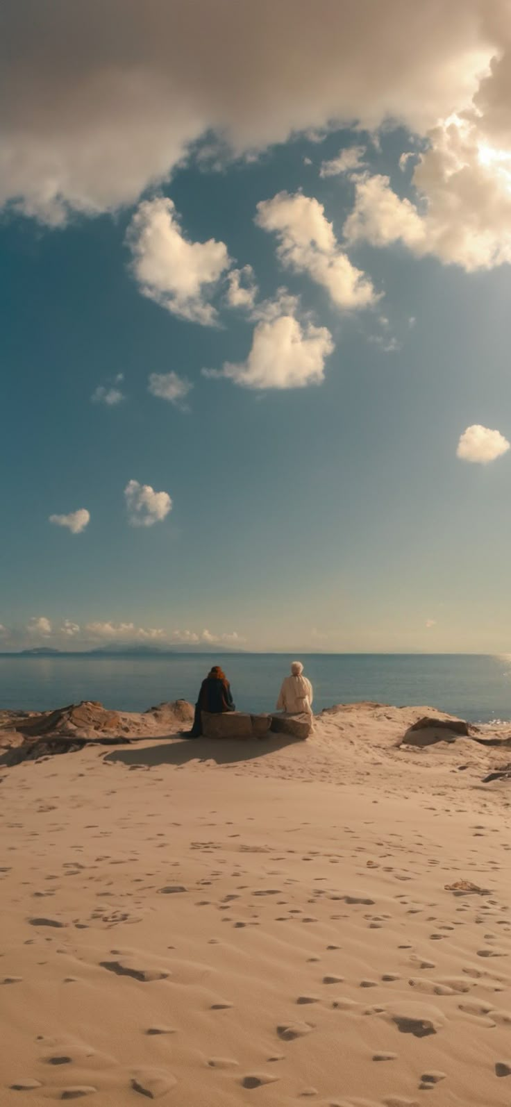 two people sitting on top of a sandy beach under a blue sky with white clouds