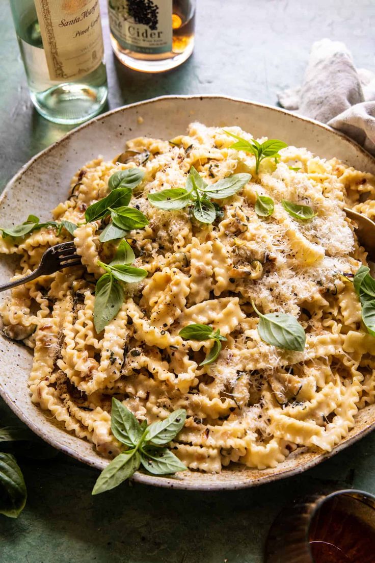 a bowl filled with pasta and basil on top of a table next to two bottles of wine