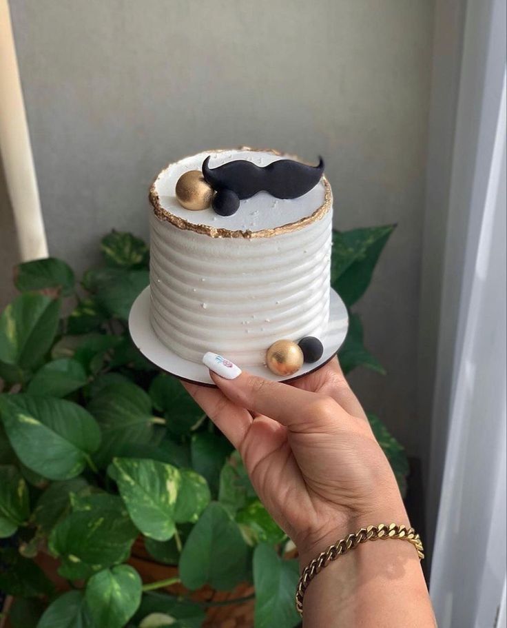 a person holding a cake with two black mustaches on top and gold balls in the middle