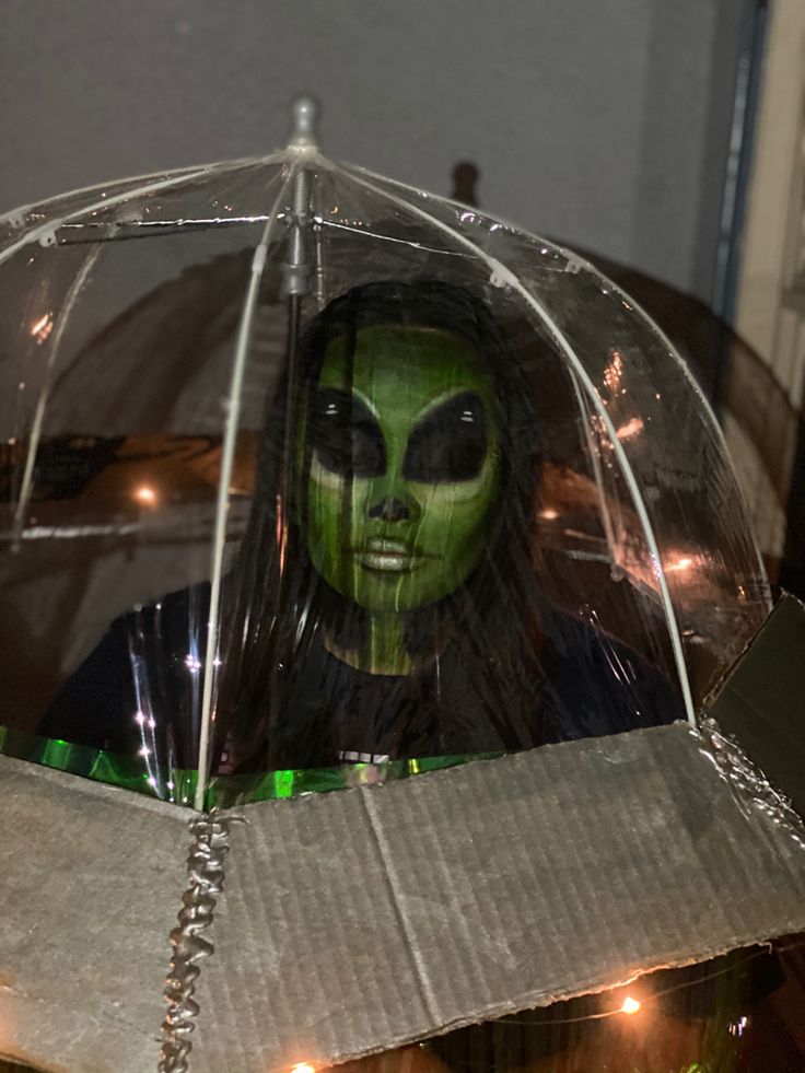 a person wearing green makeup and holding an open clear umbrella with lights in the background