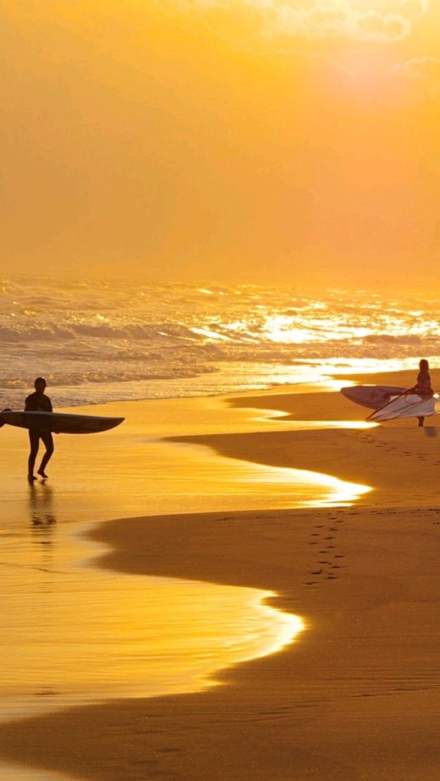 two people carrying surfboards on the beach at sunset