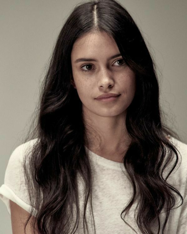 a woman with long dark hair wearing a white shirt and looking at the camera while standing in front of a gray background