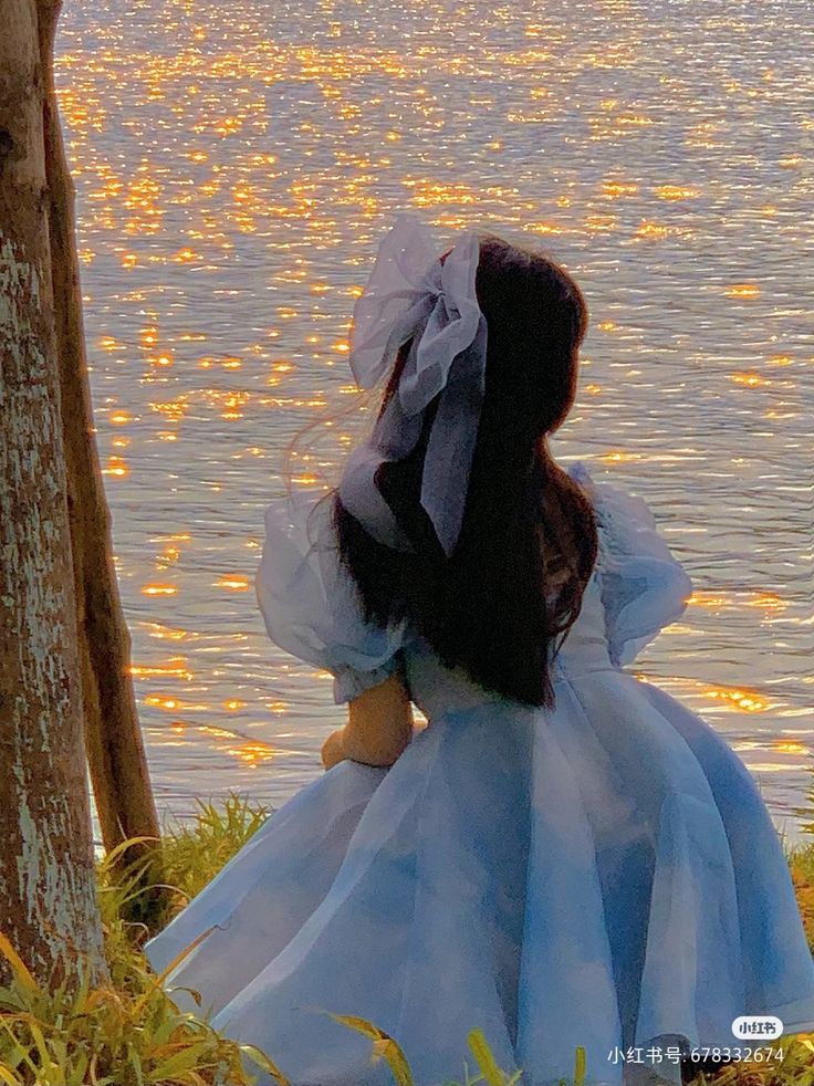 a woman in a blue dress sitting next to a tree looking out at the water