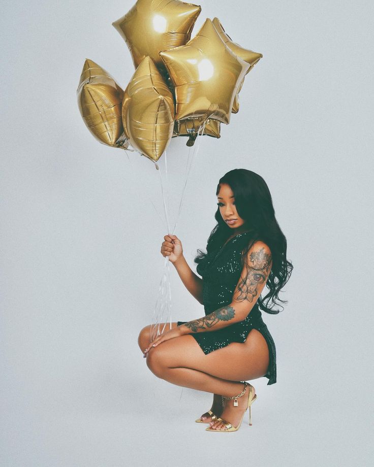 a woman holding some gold balloons in her hand and sitting on the ground with it's legs crossed