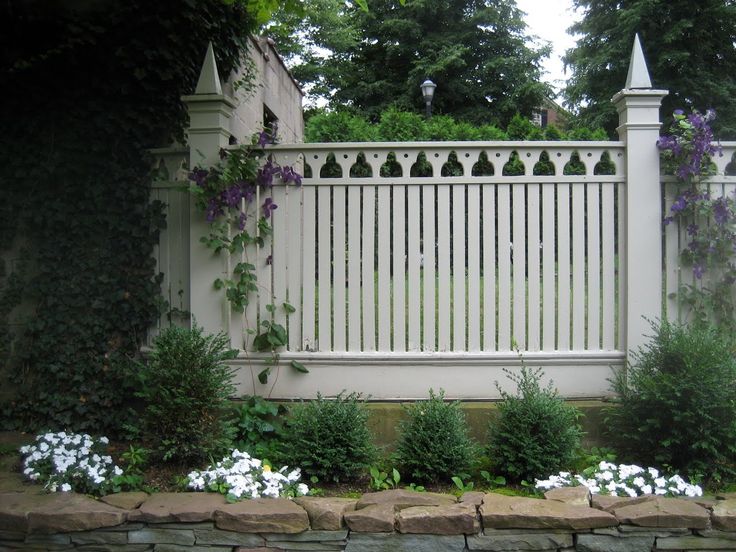 a white picket fence surrounded by flowers and greenery in front of a stone wall