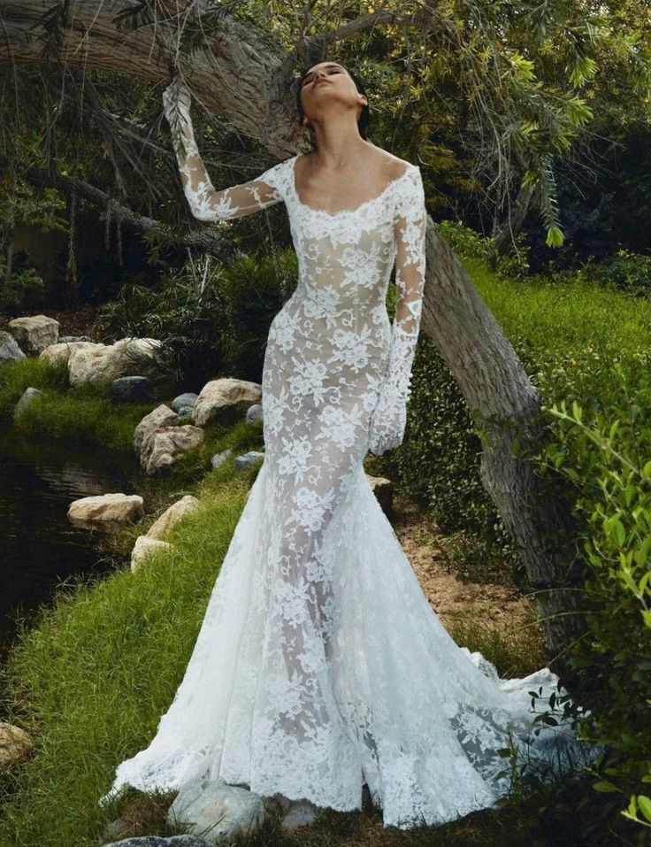 a woman in a white wedding dress is standing by some trees and grass with her hands on her head