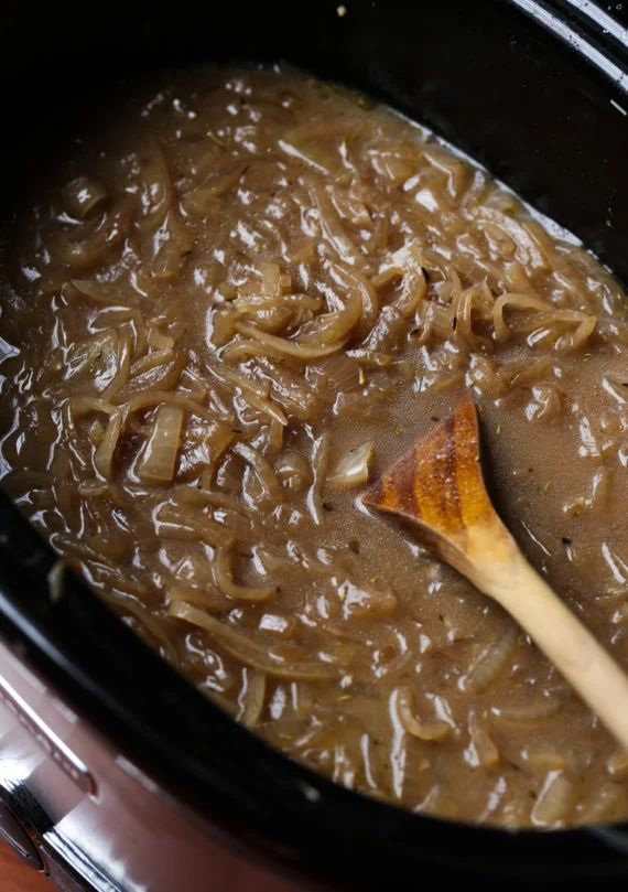 a wooden spoon in a slow cooker filled with brown liquid and onion gravy