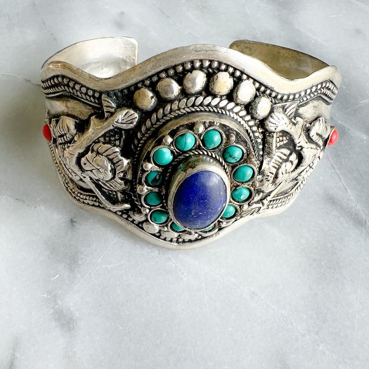 Silver tribal Aztec design bracelet with lapis lazuli set semi-precious stone. Features turquoise and coral inlays. Adjustable, can squeeze smaller or larger to fit. Comes in linen travel pouch. About 3.5” width. Open cuff style bangle. Made in USA. Artisan Blue Bangle Cuff Bracelet, Artisan Blue Cuff Bangle Bracelet, Artisan Handmade Blue Bangle, Adjustable Artisan Blue Bangle, Artisan Blue Cuff Bracelet For Festivals, Unique Blue Cuff Bracelet For Festival, Unique Blue Nickel-free Cuff Bracelet, Bohemian Lapis Lazuli Gemstone Bracelets, Traditional Blue Gemstone Bracelets