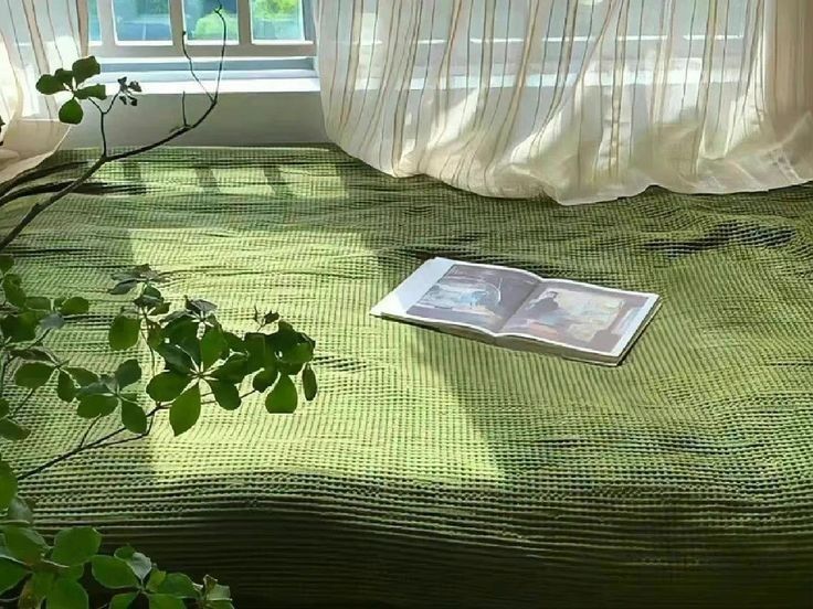 an open book laying on top of a bed next to a plant in front of a window