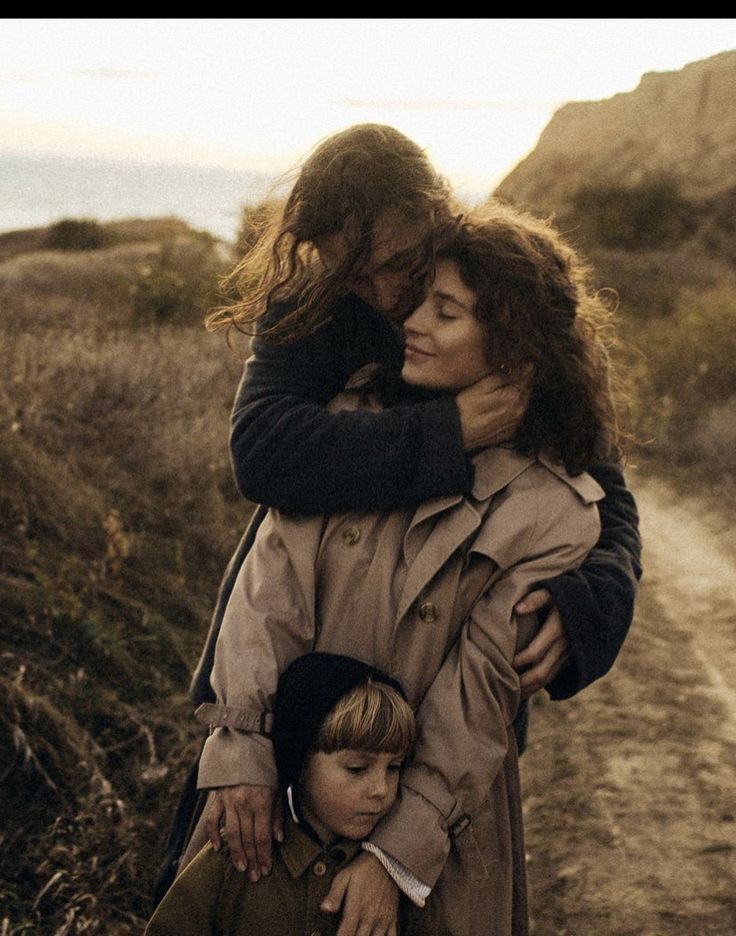 two women and a boy hugging each other on a dirt path near the ocean with mountains in the background