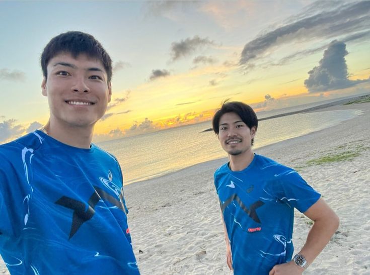 two men standing on the beach at sunset with one wearing a blue t - shirt