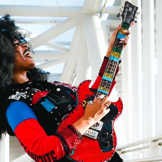 a woman with black hair holding a red and blue guitar in her right hand as she poses for the camera