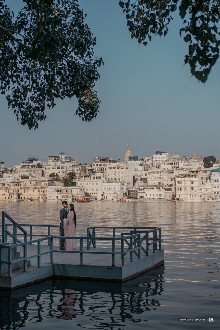 a bride and groom standing on a dock in front of the water with cityscape in the background